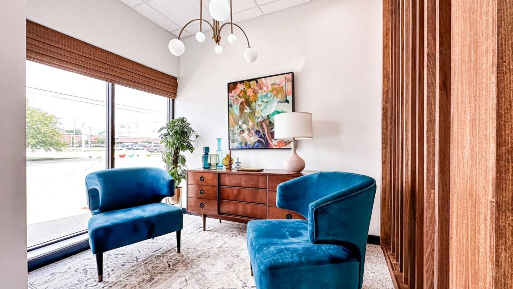 Office lounge with blue velvet chairs, floral artwork, and wood accent divider.