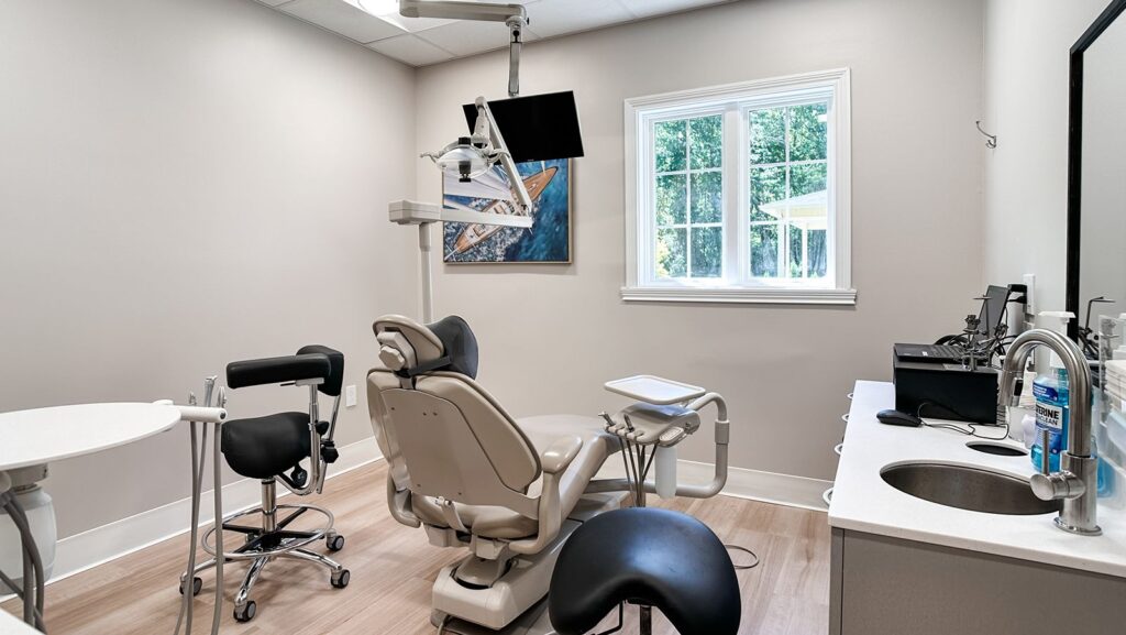 Dental exam room with treatment chair, wall-mounted monitor, and large window.